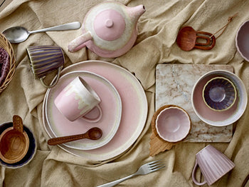 A flat lay of pink ceramic tableware and wooden spoons rests on a beige cloth, featuring the Bloomingville Gabrielle Trivet by Bloomingville for table protection. The pastel pieces have a handmade look, complemented by a fork and small basket.