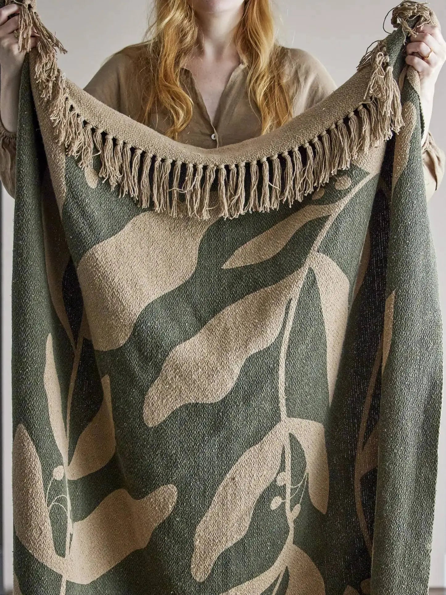A person with long, wavy blonde hair holds up the Bloomingville Geneva Throw by Bloomingville, a woven recycled cotton blanket with beige fringe and a cozy green botanical leaf pattern. The person wears a beige shirt.