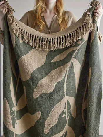 A person with long, wavy blonde hair holds up the Bloomingville Geneva Throw by Bloomingville, a woven recycled cotton blanket with beige fringe and a cozy green botanical leaf pattern. The person wears a beige shirt.