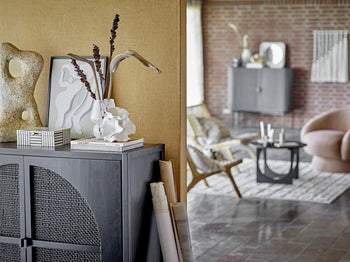 A modern living space featuring a grey cabinet, abstract sculptures, books, dried plants in the Bloomingville Halim Vase by Bloomingville. Wicker furniture, a round armchair, and stylish decor enhance the room’s charm against a brick wall.
