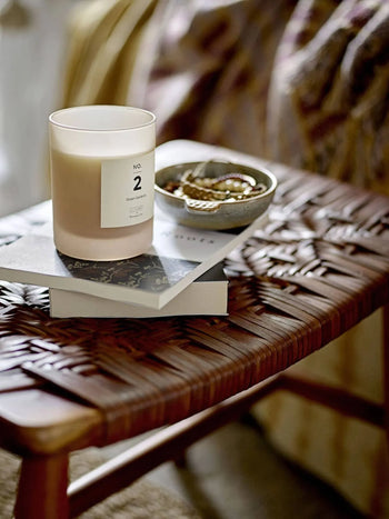 A white candle labeled “No. 2” sits on stacked books atop a woven rattan side table. Next to it, jewelry rests in the Bloomingville Hariet Bowl by Bloomingville, with patterned fabric visible in the background.