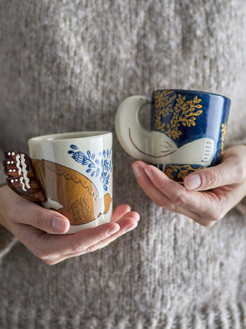 A person in a beige knit sweater holds two Bloomingville Hezha Cups—stoneware mugs by Bloomingville, each hand-decorated with bird motifs and intricate blue and brown patterns: one mainly white, the other deep blue.