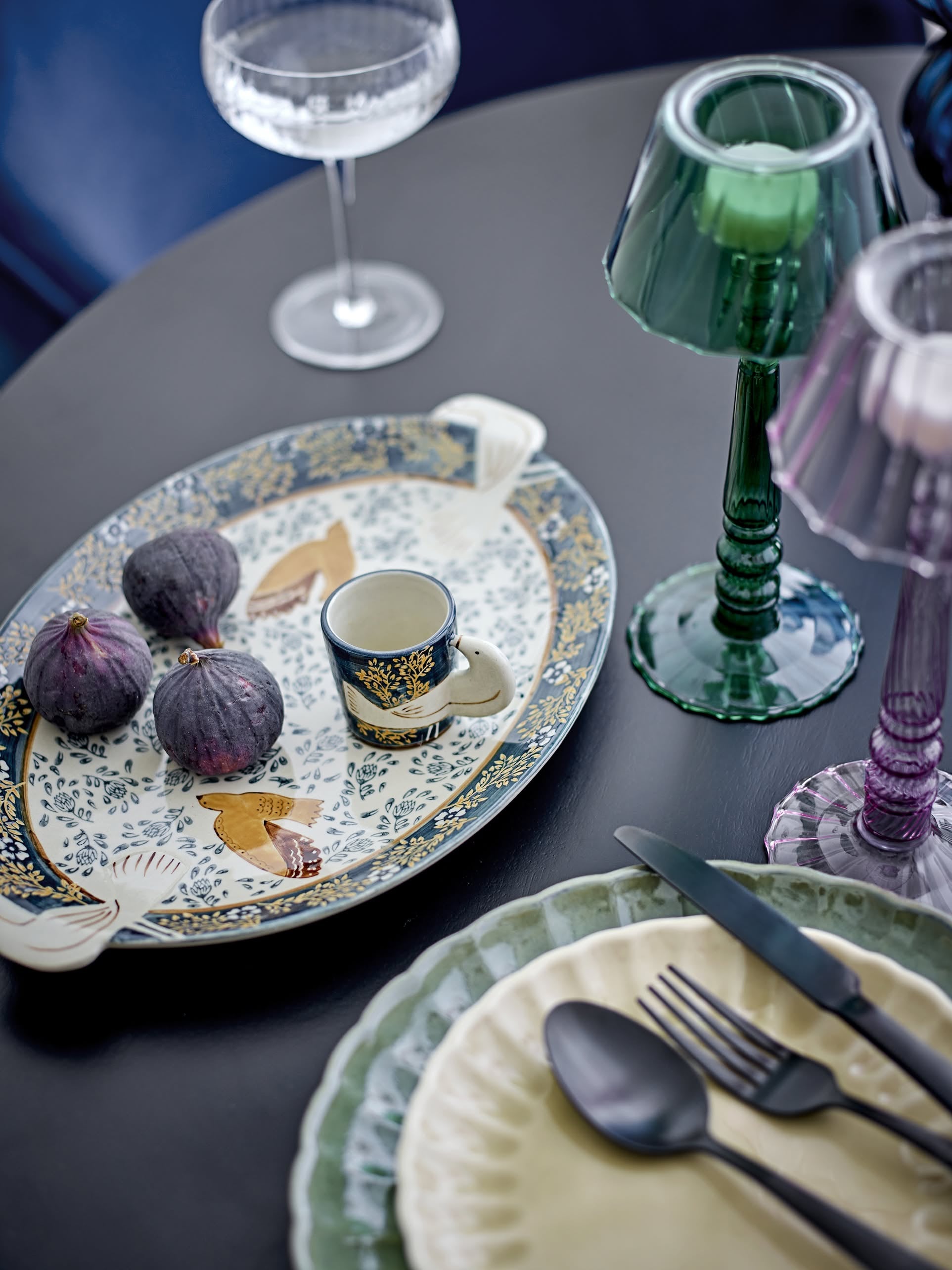 A decorative table setting features a hand-decorated bird motif platter with figs, the Bloomingville Hezha Cup - Blue, colored glass candle holders, a green-rimmed plate, cutlery, and a wine glass on a dark table.
