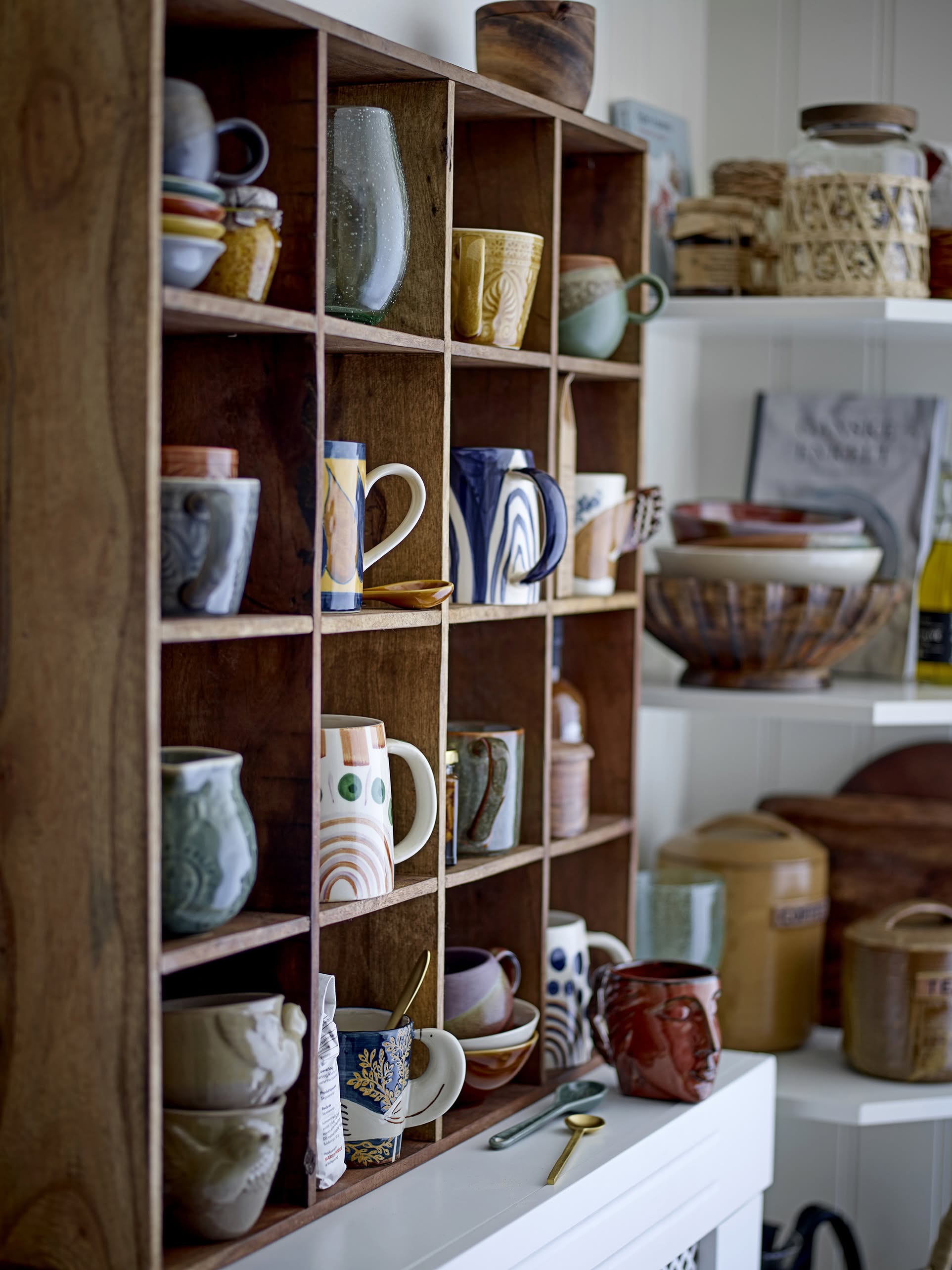 A wooden cubby shelf displays colorful ceramic mugs, including the Bloomingville Hezha Cup - Blue; nearby shelves feature other stoneware bowls, jars, and kitchen items in a cozy, eclectic kitchen setting.