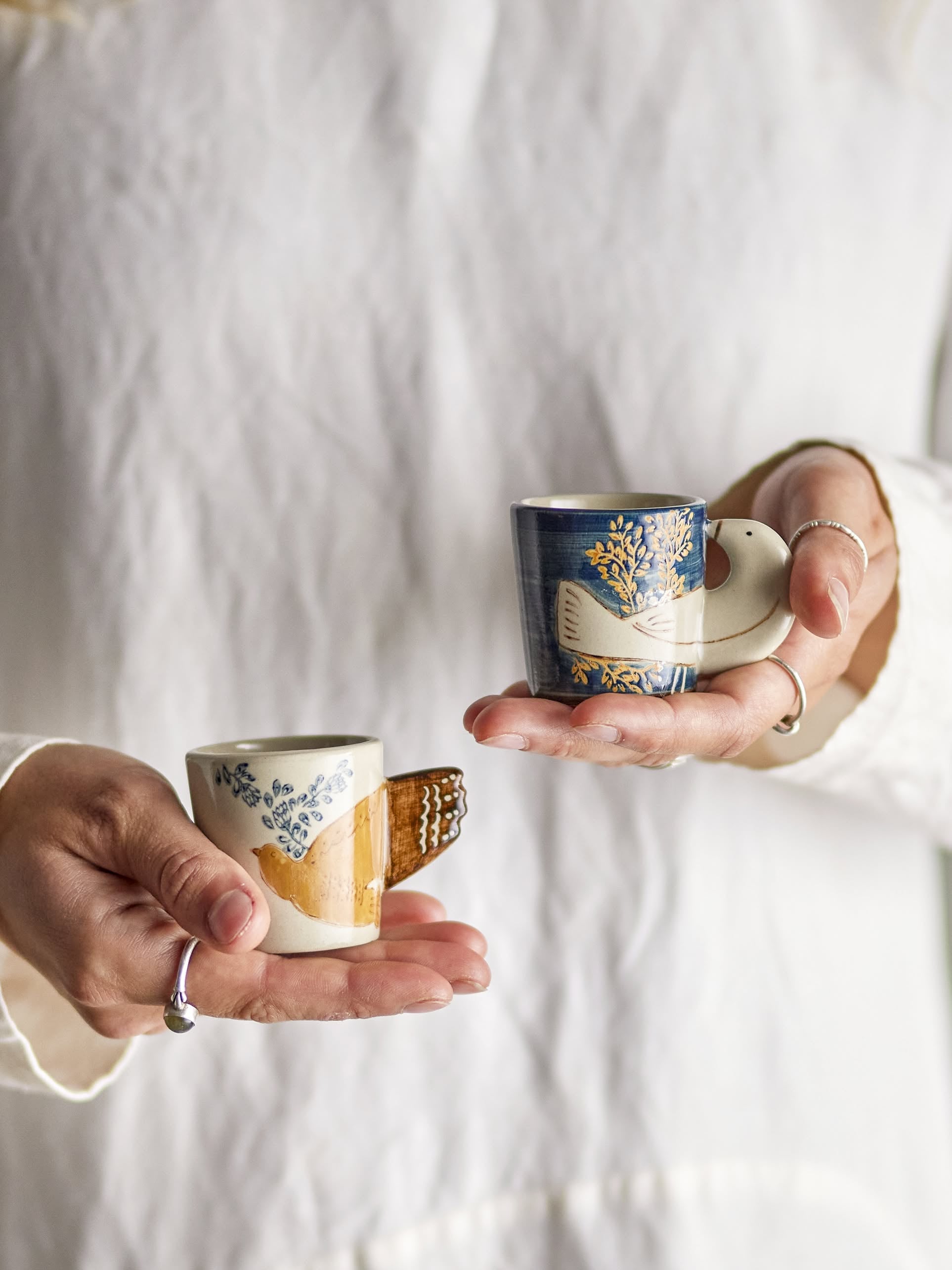 A person in a white shirt holds two Bloomingville Hezha Cups—one blue with a tree and bird motif, and one cream-colored—both featuring bird wing handles.