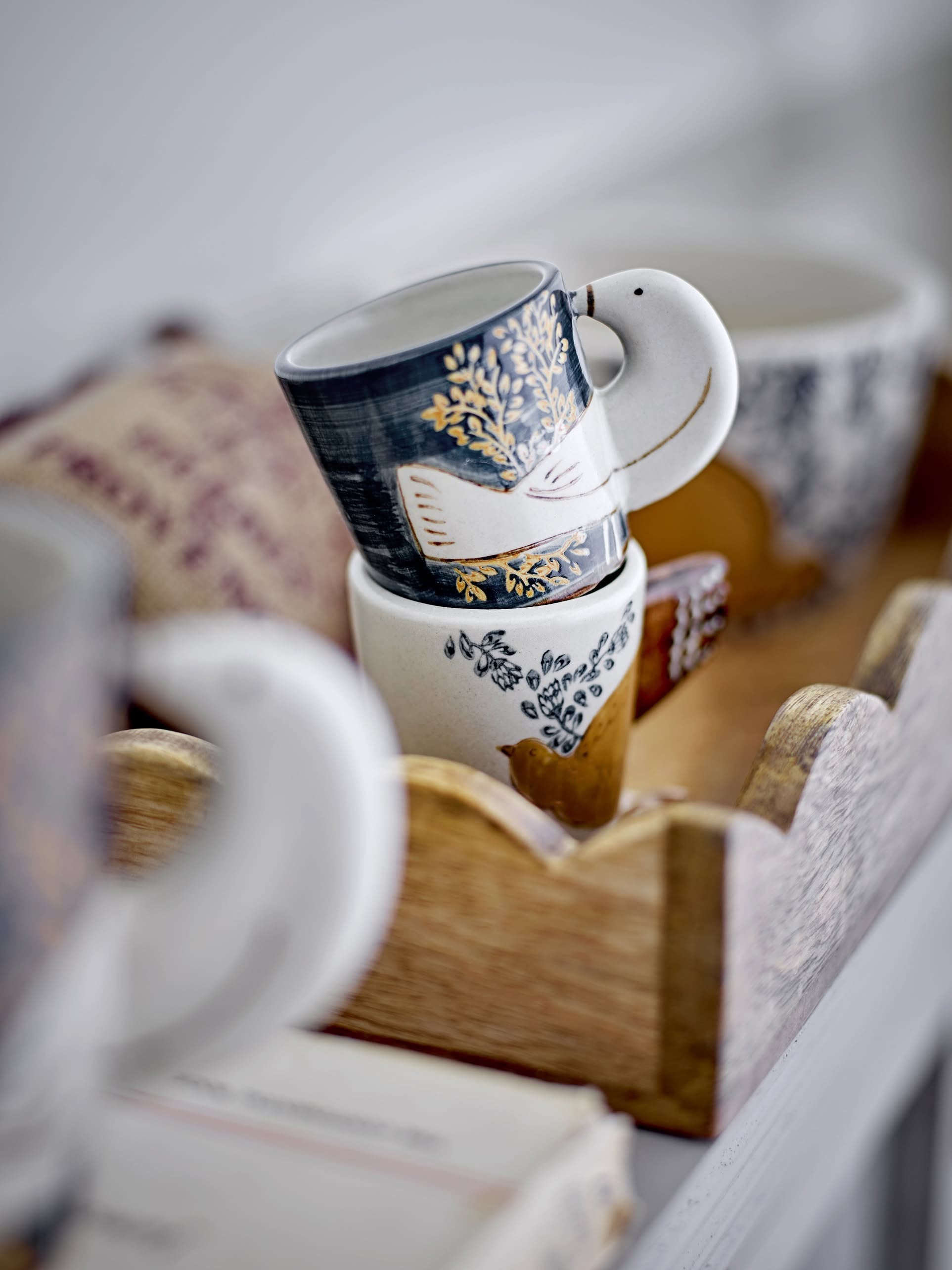 The Bloomingville Hezha Cups by Bloomingville, featuring charming bird and foliage designs, are stacked in a wooden tray with softly blurred background objects.