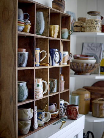 A wooden cubby shelf displays handmade mugs and cups, featuring the Bloomingville Hezha Cup - Nature with a bird motif by Bloomingville, surrounded by shelves of stoneware bowls, jars, and rustic pottery in a cozy kitchen setting.