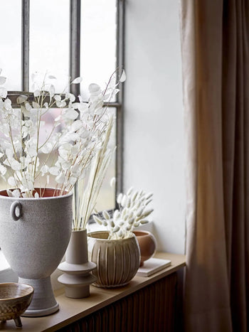 A windowsill featuring the Bloomingville Isold Vase by Bloomingville, filled with white dried flowers and grasses next to a closed beige curtain, creates a rustic look as soft natural light pours in.