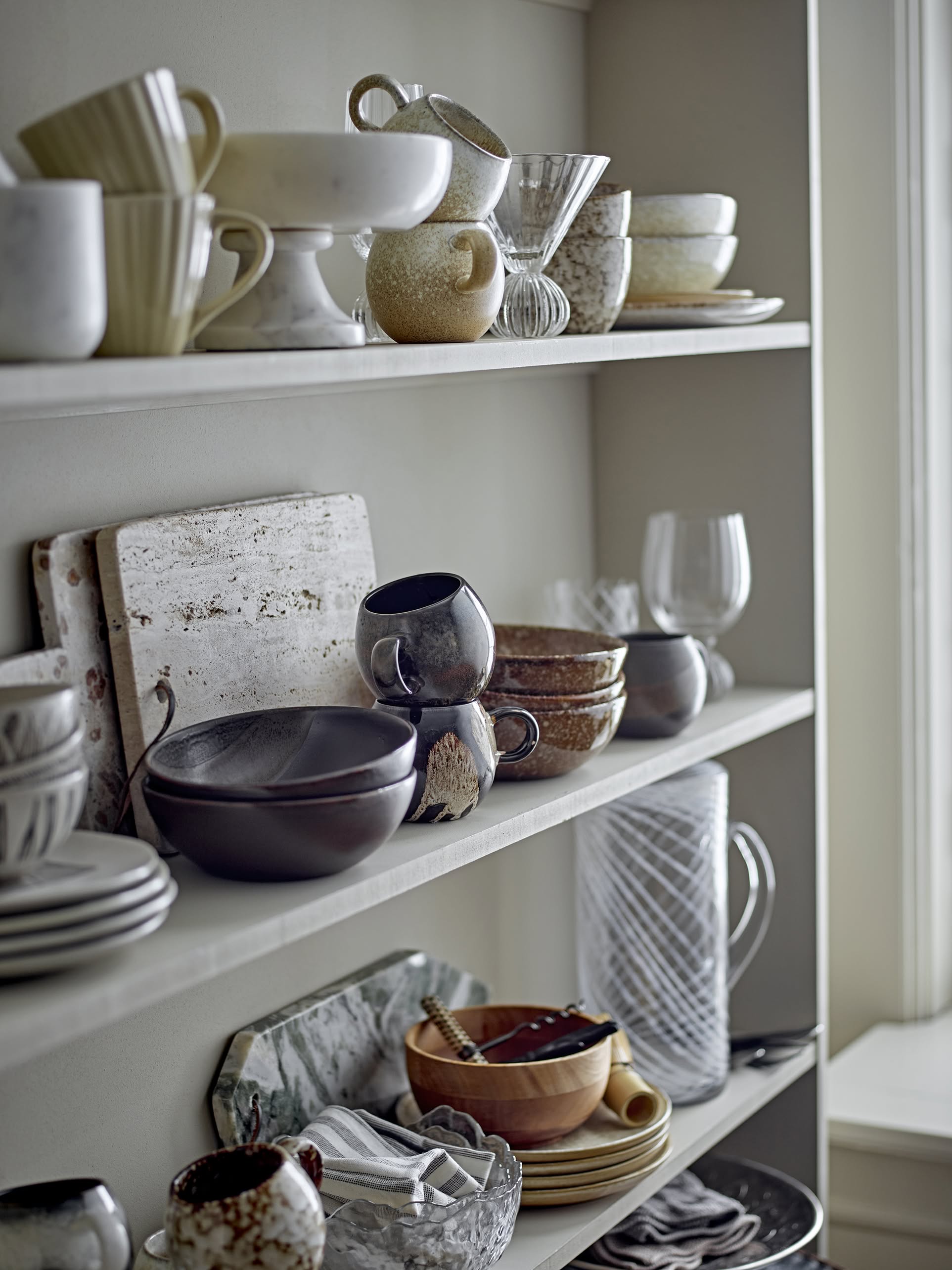Open shelves display assorted ceramic mugs, bowls, plates, glassware, and the Bloomingville Izabel Cutting Board by Bloomingville with natural veining, all arranged neatly as a decorative kitchen accent in neutral earthy tones.