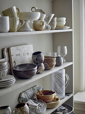 Open shelves display assorted ceramic mugs, bowls, plates, glassware, and the Bloomingville Izabel Cutting Board by Bloomingville with natural veining, all arranged neatly as a decorative kitchen accent in neutral earthy tones.