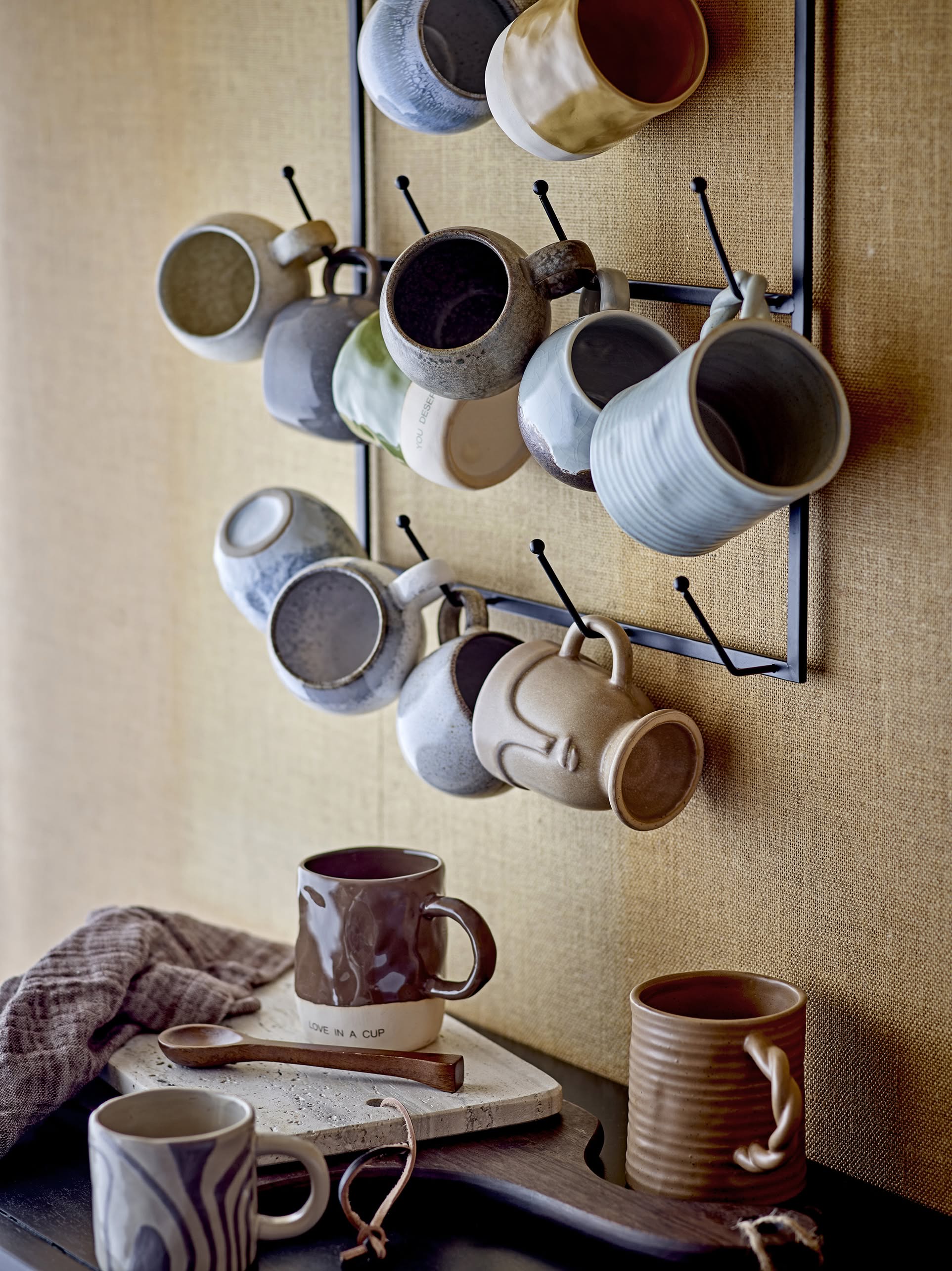 A metal rack holds assorted ceramic mugs on a beige wall. Below, two mugs rest on the Bloomingville Izabel Cutting Board by Bloomingville, showcasing natural veining, next to a brown-striped cloth and wooden spoon.