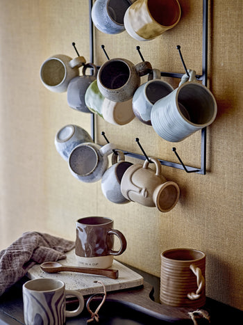 A metal rack holds assorted ceramic mugs on a beige wall. Below, two mugs rest on the Bloomingville Izabel Cutting Board by Bloomingville, showcasing natural veining, next to a brown-striped cloth and wooden spoon.