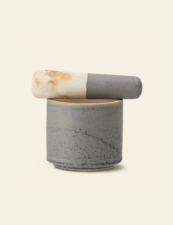 The Bloomingville Kendra Mortar & Pestle features a textured gray ceramic finish, ideal for grinding herbs and spices. The pestle rests atop the mortar, which is displayed on a light beige background.