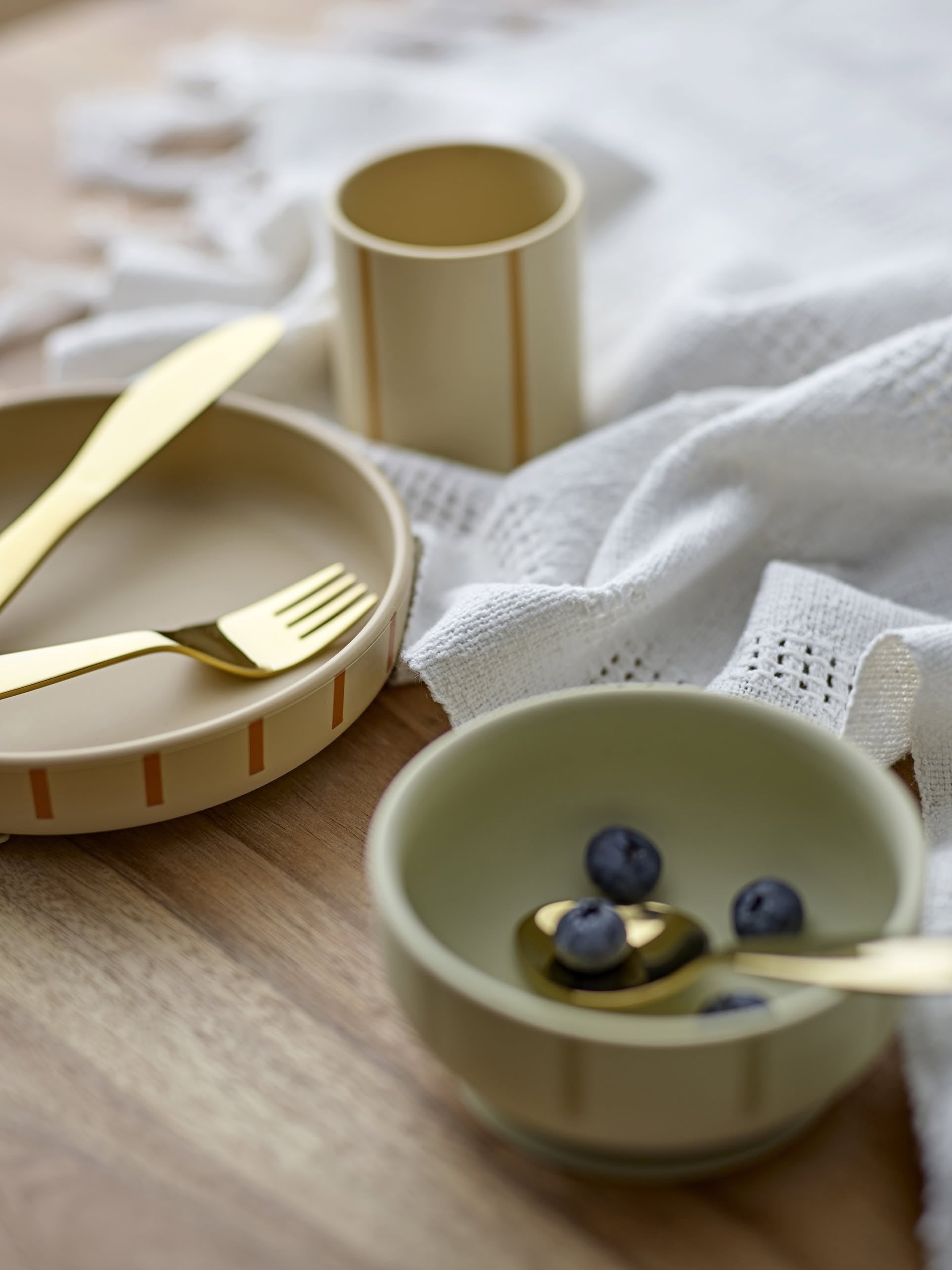 The Bloomingville Levi Tableware Set features two ceramic bowls—one with blueberries and a gold spoon, the other with a gold fork and knife—on a wooden table near a white textured cloth and beige cup. Perfect gift for young children.