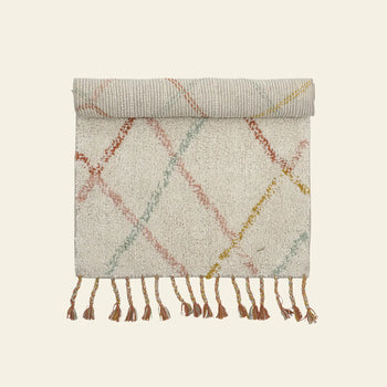 The cream Bloomingville Lilou Rug features pastel pink, yellow, and red diagonal lines, is rolled at the top with tassels along the bottom edge—ideal for adding color to a child’s room.