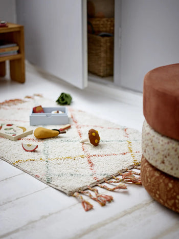A cozy Bloomingville Lilou Rug with tassels rests on a white floor, surrounded by wooden toy food, a small toy box, a round cushion seat, wooden shelf, and woven basket in an open closet—completing this charming children’s room.