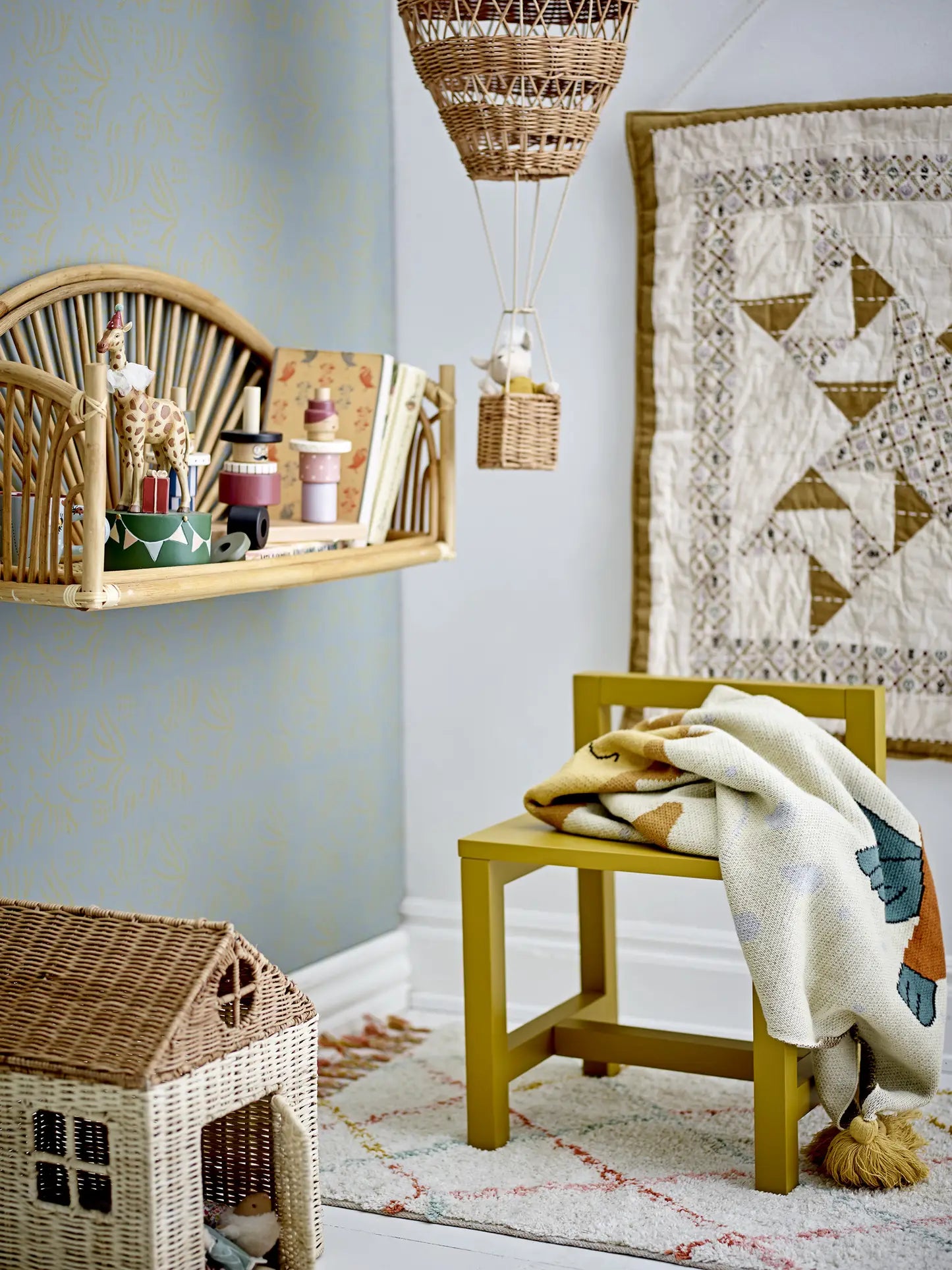 A cozy children’s room features a yellow chair with a patterned blanket, the Bloomingville Lilou Rug by Bloomingville, a wicker dollhouse, wooden shelves with toys and books, a quilted wall hanging, and a woven hot air balloon decoration.