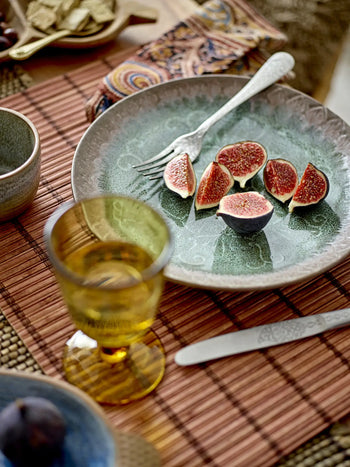A green patterned plate with sliced figs and a fork sits on the Bloomingville Maggi Placemat in brown. Nearby, a yellow glass, knife, cup, and patterned napkin complete the inviting table setup by Bloomingville.