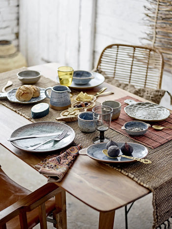 A rustic wooden table set for a meal with ceramic plates, cups, figs, bread, olives, cheese, wine glasses, and the Bloomingville Maggi Placemat in brown by Bloomingville. Two chairs flank the table—one wicker and one wooden.
