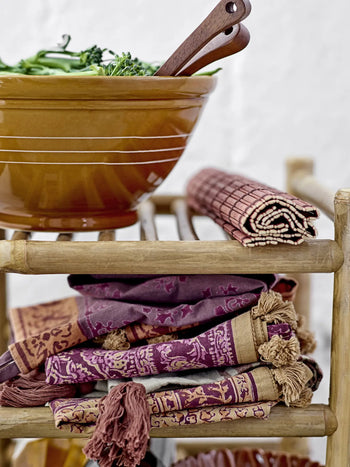 The Bloomingville Maggi Placemat - Brown by Bloomingville features a wooden rack holding a brown ceramic bowl, utensils, a red and beige napkin, and purple and gold textiles, creating striking table decor.