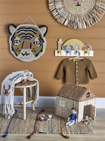 A cozy, animal-themed children’s room with a tiger wall hanging, Bloomingville Mallen Coat Rack by Bloomingville, a brown knit cardigan, wicker stool with blanket, toy house, and two dolls on a woven rug.