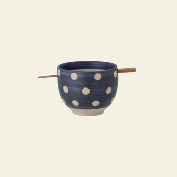 The Bloomingville Masami Bowl w/Chopsticks by Bloomingville is a blue stoneware bowl with white polka dots, featuring chopsticks resting across the top. This stylish kitchenware is displayed against a plain, light background.
