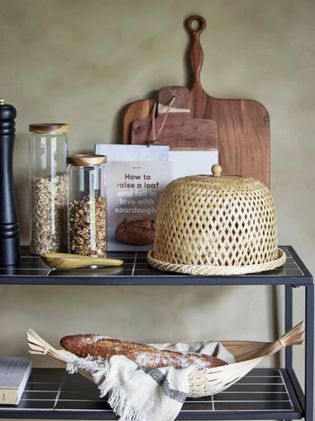 A kitchen shelf holds two jars of grains, a pepper grinder, the Bloomingville Masami Spoon, two cutting boards, How to Raise a Loaf and Fall in Love with Sourdough, a wicker food cover, and a loaf of bread—perfect cooking inspiration.