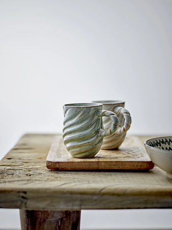Two Bloomingville Miriam Mugs with textured twisted and spiral designs sit on a wooden board on a rustic table, next to a small bowl featuring a green glaze zigzag rim, all set against a plain, light background.
