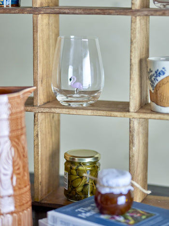 A wooden shelf holds a Bloomingville Misa Drinking Glass by Bloomingville with a tiny flamingo inside, a jar of pickles, a ceramic pitcher, a mug, a small jar with a cloth cover, and some books.