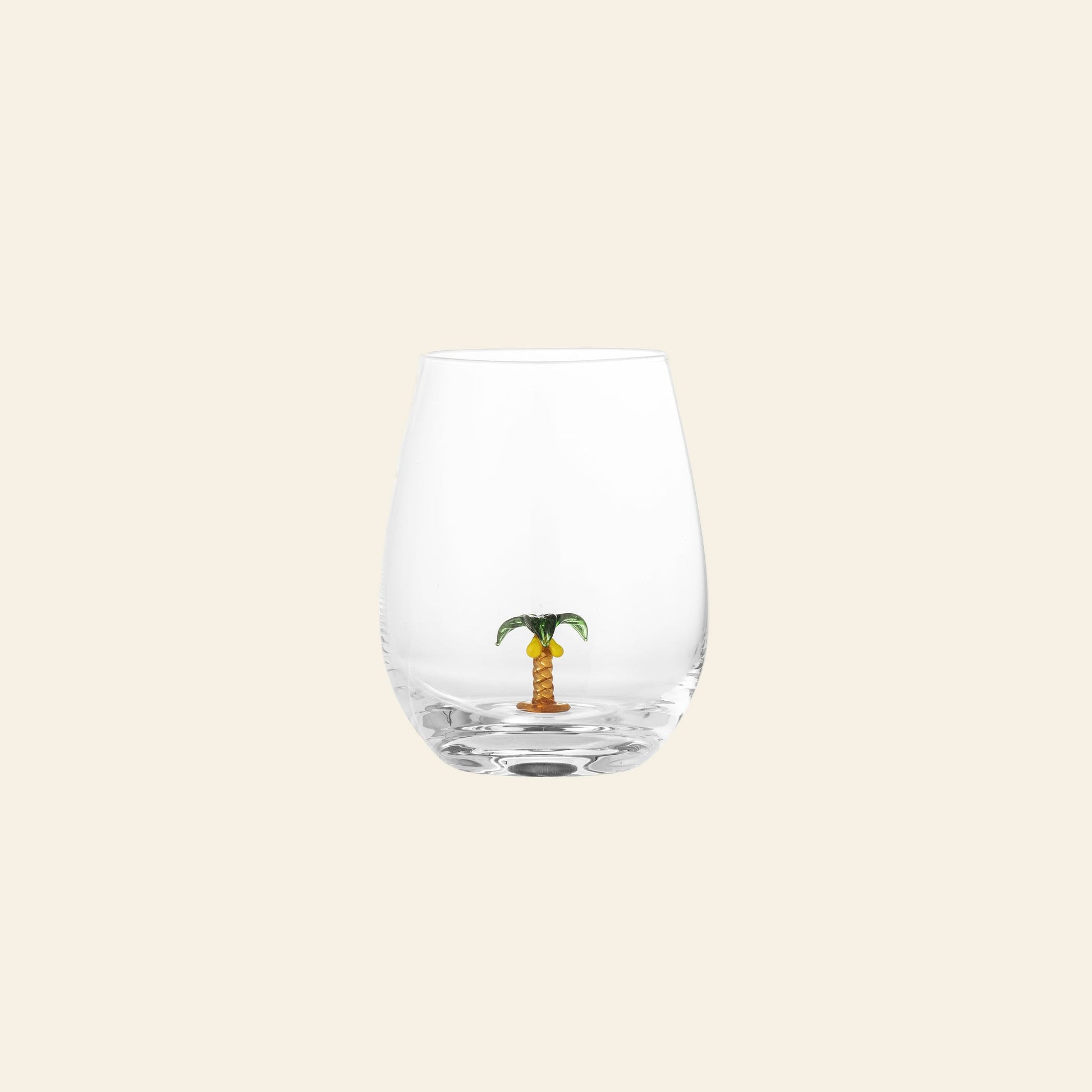 The Bloomingville Misa Drinking Glass by Bloomingville is a unique handmade glass with a small decorative palm tree figurine at the bottom, set against a plain light beige background.
