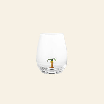 The Bloomingville Misa Drinking Glass by Bloomingville is a unique handmade glass with a small decorative palm tree figurine at the bottom, set against a plain light beige background.