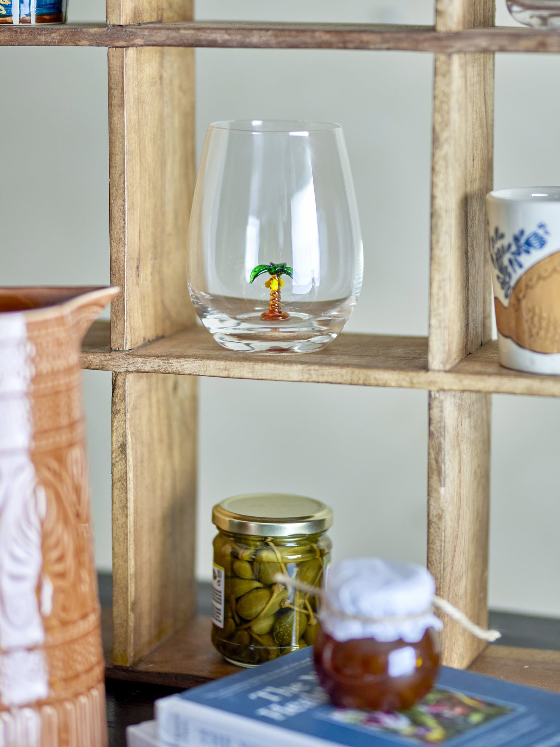 A wooden shelf displays the Bloomingville Misa Drinking Glass with a small palm tree inside, a ceramic mug, a jar of olives, a fabric-covered jar, books, and a large brown pitcher in the foreground.
