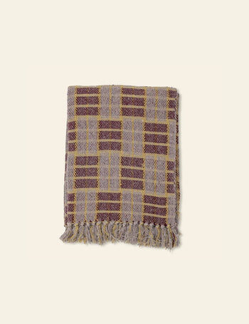 The Bloomingville Mody Throw by Bloomingville is a folded woven blanket with a plaid pattern in warm yellow, purple, and beige hues, natural earthy accents, and fringe on the short ends, set against a plain light background.