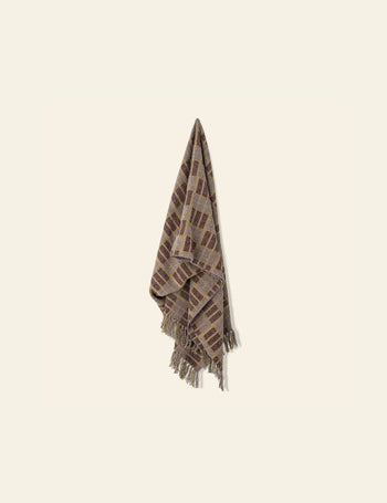 The Bloomingville Mody Throw by Bloomingville, featuring a brown and beige geometric checkered pattern with fringed edges, hangs on a light cream wall with its bottom corners draped loosely downward.