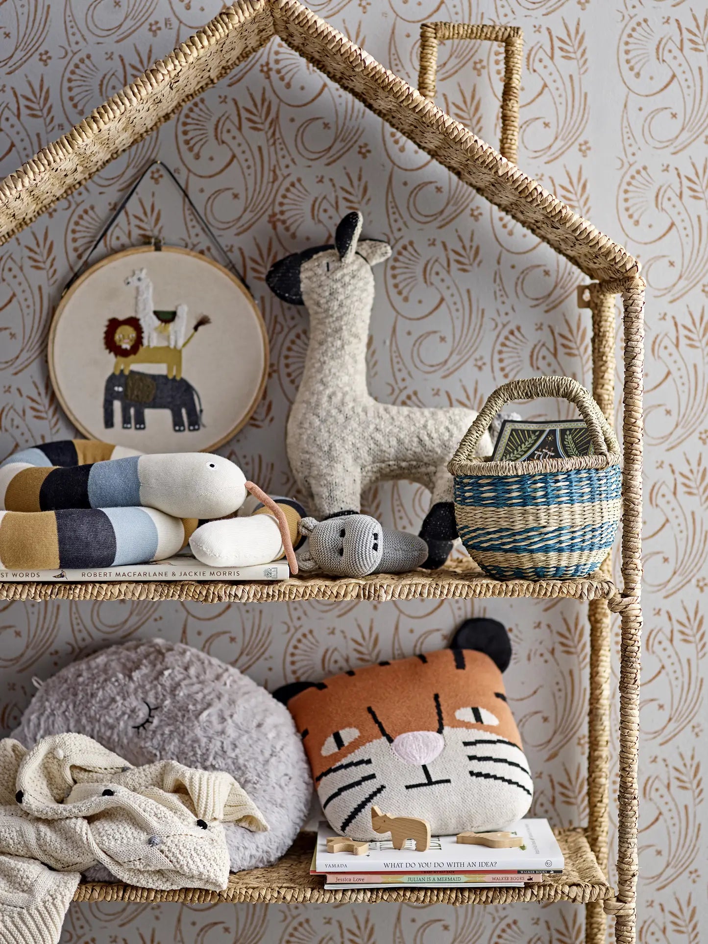 A woven shelf holds a Bloomingville llama hoop art, plush llama, striped snake toy, hippo toy, blue basket, gray plushie, Bloomingville Muzamel Cushion as a decorative accent, folded blankets, and children’s books against patterned wallpaper.
