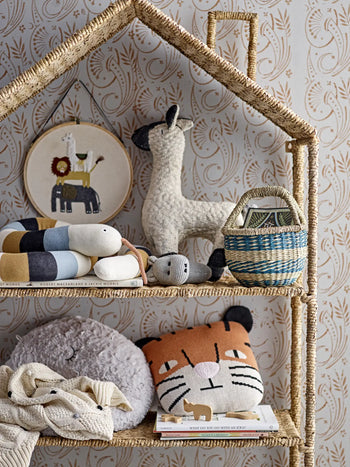 A woven shelf holds a Bloomingville llama hoop art, plush llama, striped snake toy, hippo toy, blue basket, gray plushie, Bloomingville Muzamel Cushion as a decorative accent, folded blankets, and children’s books against patterned wallpaper.