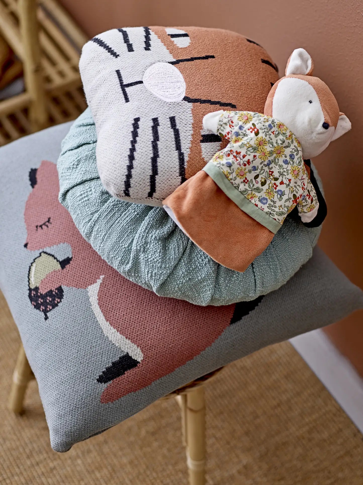A wooden chair by a peach-colored wall is decorated with a stack of animal-themed pillows, including the Bloomingville Muzamel Cushion and one with a lion face, topped by a plush fox doll in a floral dress for a whimsical accent.