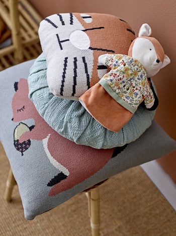 A wooden chair by a peach-colored wall is decorated with a stack of animal-themed pillows, including the Bloomingville Muzamel Cushion and one with a lion face, topped by a plush fox doll in a floral dress for a whimsical accent.