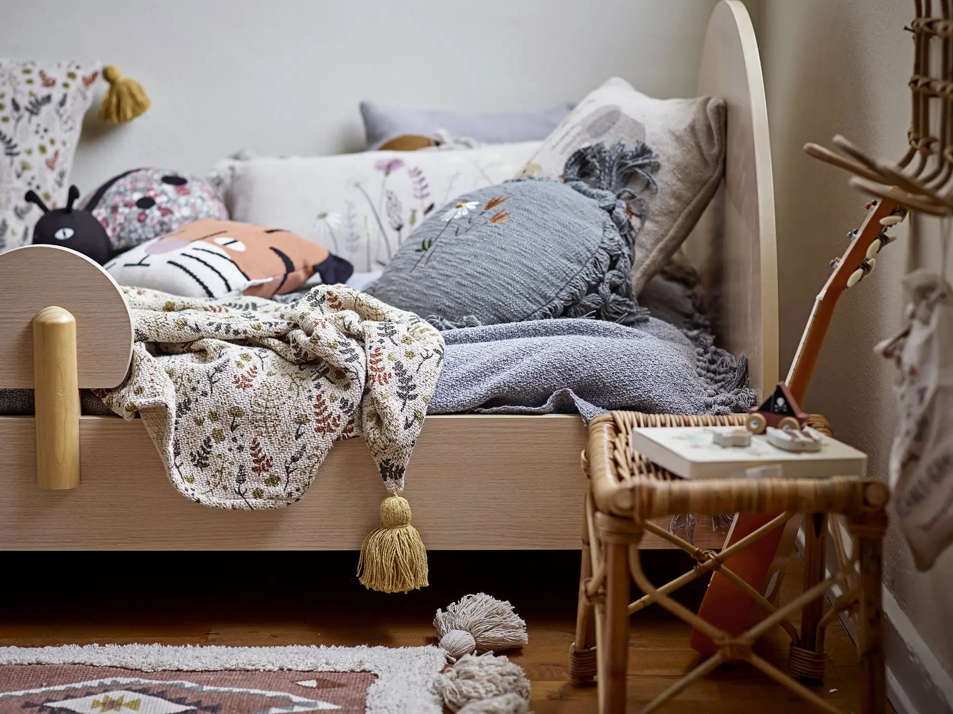 A cozy childs bed is styled with floral bedding, decorative pillows, and the playful Bloomingville Muzamel Cushion by Bloomingville. A wicker stool with a guitar sits beside the bed, while a soft rug lies on the wooden floor.