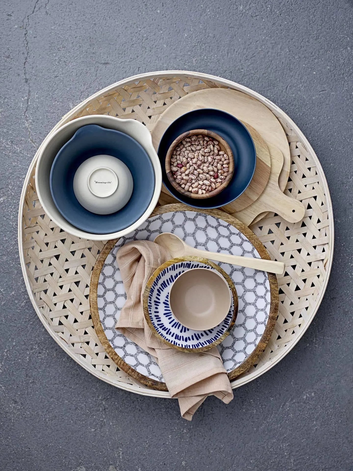 A round woven tray holds a blue mixing bowl, the Bloomingville Nolia Plate (25cm), a bowl of pinto beans on a wooden board, a beige bowl with striped rim, pale napkin, and wooden spoon—comfort foods artfully arranged.