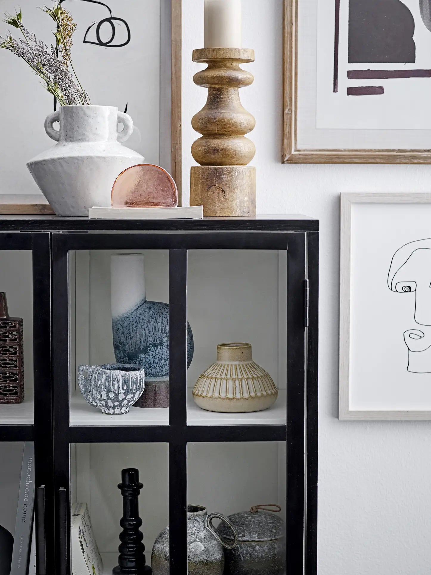 A black glass-front cabinet displays ceramic vases, bowls, and a Bloomingville Oney Flowerpot. On top are a white jug, wooden candle holder, and framed abstract art decorating the white wall behind.