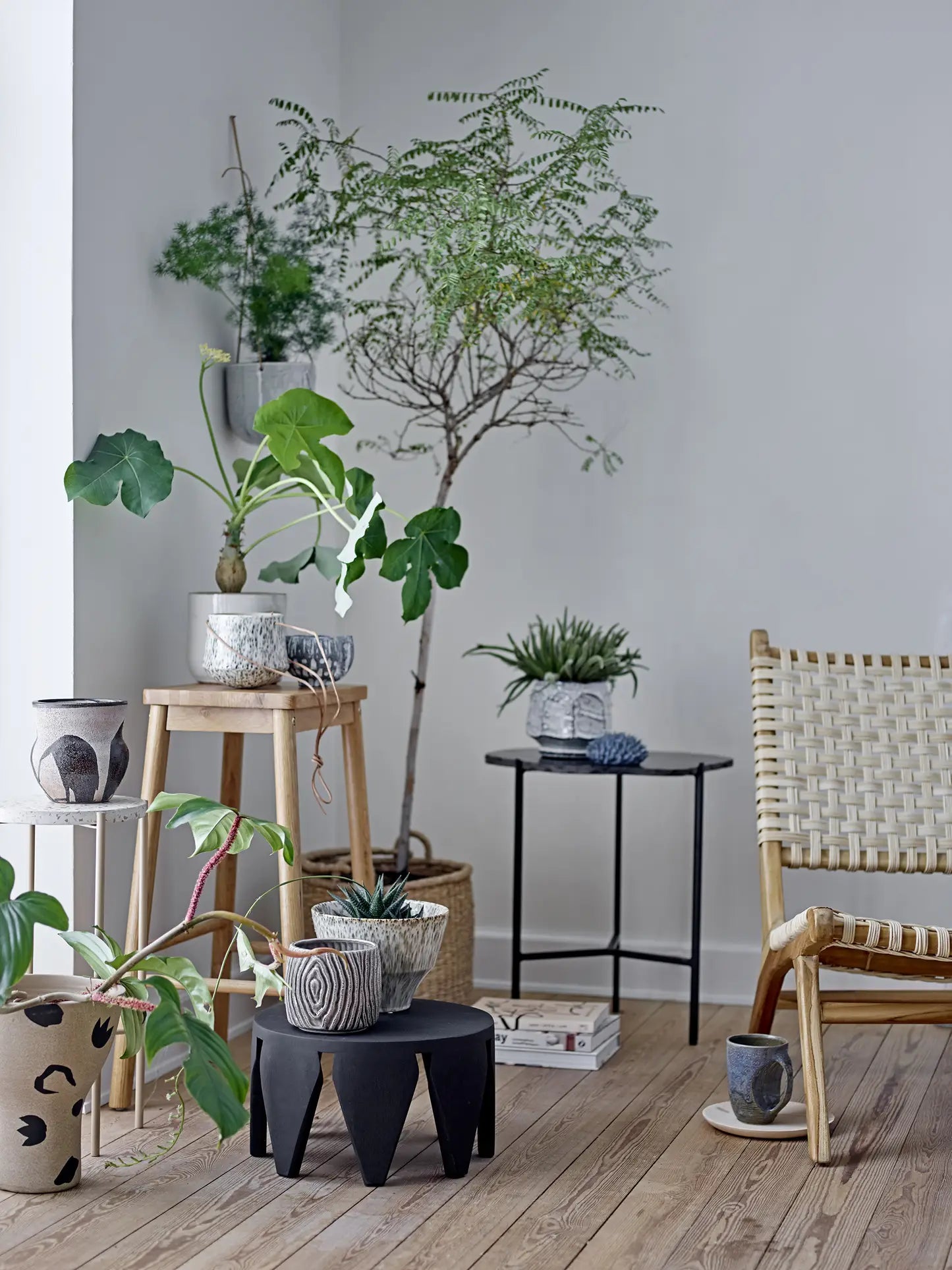 A bright, minimalist room with wooden floors, lush potted plants in Bloomingville Oney Flowerpots by Bloomingville on stools and tables, a woven chair, and a mug on a small black table, creating a cozy, natural atmosphere with handmade accents.