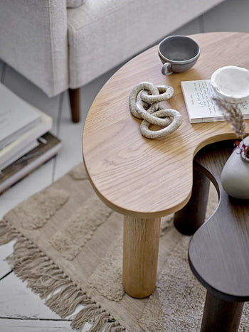 A kidney-shaped wooden coffee table with decorative objects, a Bloomingville Raben Cappuccino Cup With Saucer, and an open notebook rests on a fringed rug beside a stack of books and a light-colored sofa.