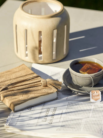 A beige candle holder, a textured tan notebook, a newspaper, and the Bloomingville Raben Cappuccino Cup With Saucer by Bloomingville sit on a sunlit light-colored outdoor table.