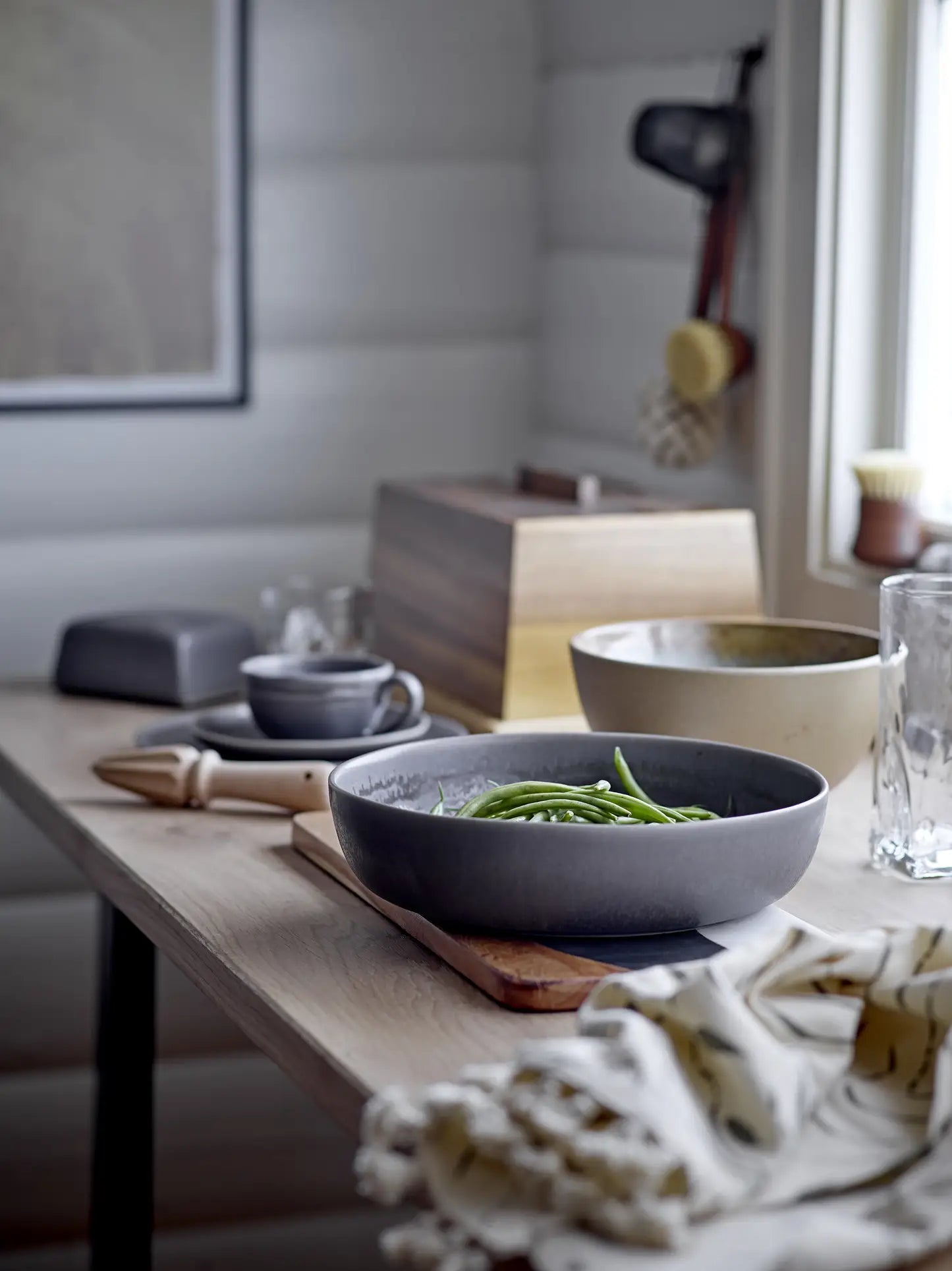 A rustic kitchen table with a Bloomingville Raben Cappuccino Cup With Saucer, a grey stoneware bowl of green beans, a glass, a wooden juicer, and other items sits near a window bathed in soft natural light.