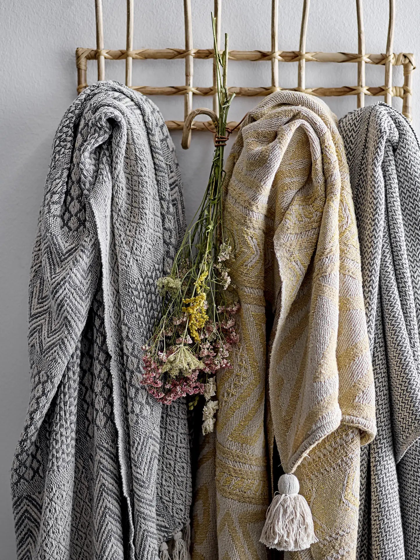 Three Bloomingville Rodion Throws in grey and beige tones hang side by side on a natural woven rack, adding decorative flair, while a small bundle of dried wildflowers is tied together and hanging between the throws.