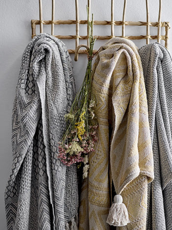 Three Bloomingville Rodion Throws in grey and beige tones hang side by side on a natural woven rack, adding decorative flair, while a small bundle of dried wildflowers is tied together and hanging between the throws.