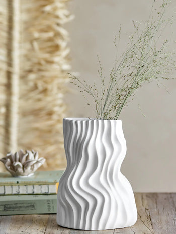 The Bloomingville Sahal Vase by Bloomingville, featuring an ocean waves texture in white ceramic, holds dried grass stems on a wooden surface beside stacked books and decor, all set against a neutral textured backdrop.