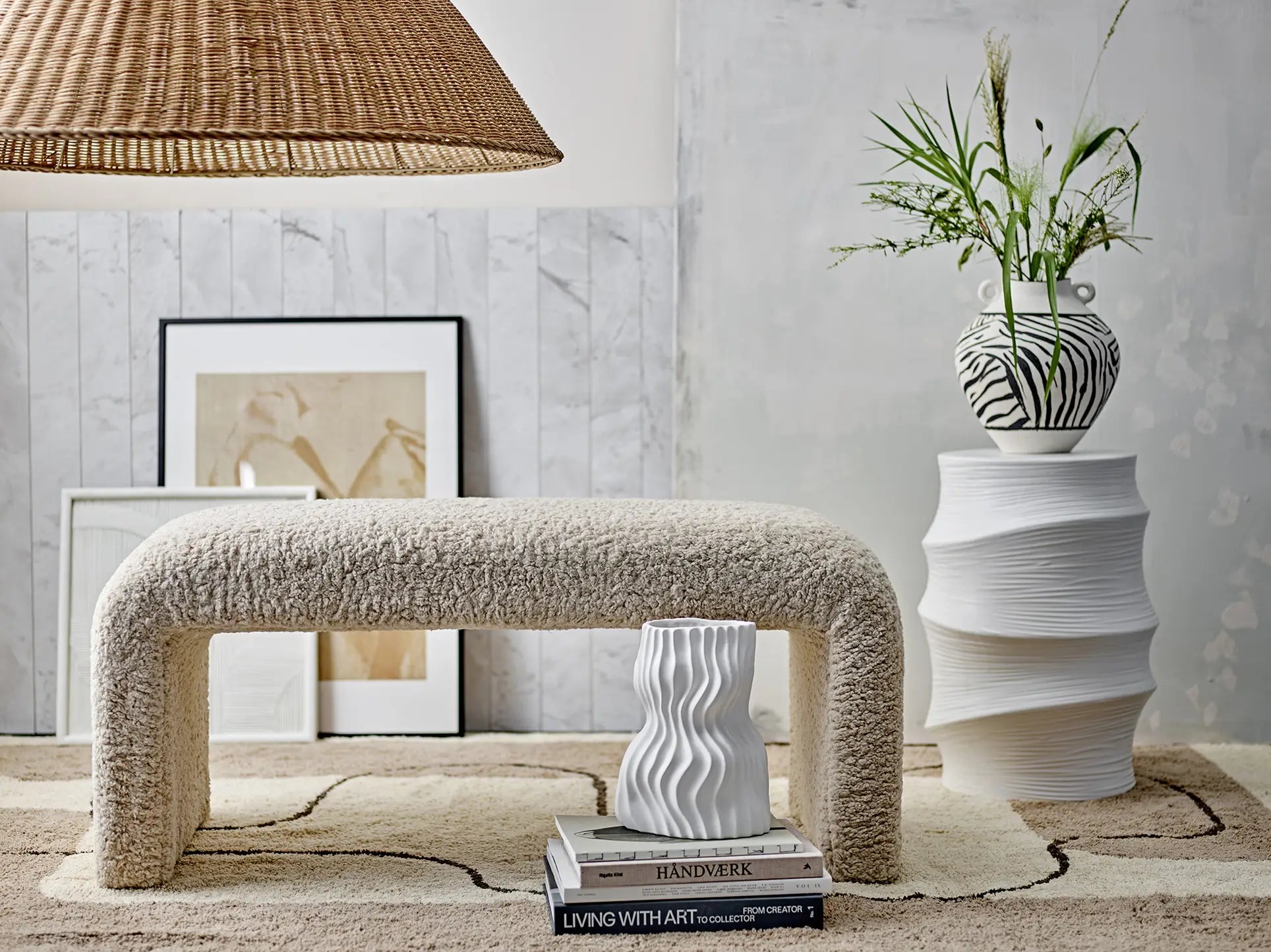 A modern, cozy room features a textured bench with books and the Bloomingville Sahal Vase, a sculptural side table with a zebra-striped decor and flowers, neutral wall art, and a woven lampshade above a patterned rug.