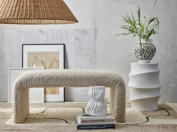 A modern, cozy room features a textured bench with books and the Bloomingville Sahal Vase, a sculptural side table with a zebra-striped decor and flowers, neutral wall art, and a woven lampshade above a patterned rug.